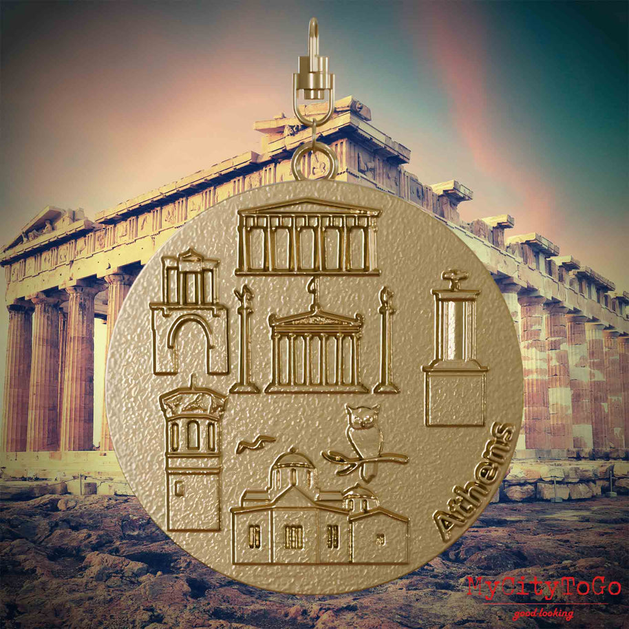 Halskette mit Drehwirbel und Rückseite eines Schmuckanhängers aus vergoldetem Edelstahl, auf dem Reliefs bekannter Sehenswürdigkeiten der Stadt Athen abgebildet sind. Den Hintergrund bildet eine Ansicht der Stadt Athen.
