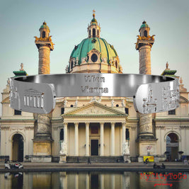 Innenseite eines silbernen Armreifs mit Reliefs bekannter Sehenswürdigkeiten der Stadt Wien und einer Gravur des Stadtnamens . Den Hintergrund bildet eine Ansicht der Stadt Wien.