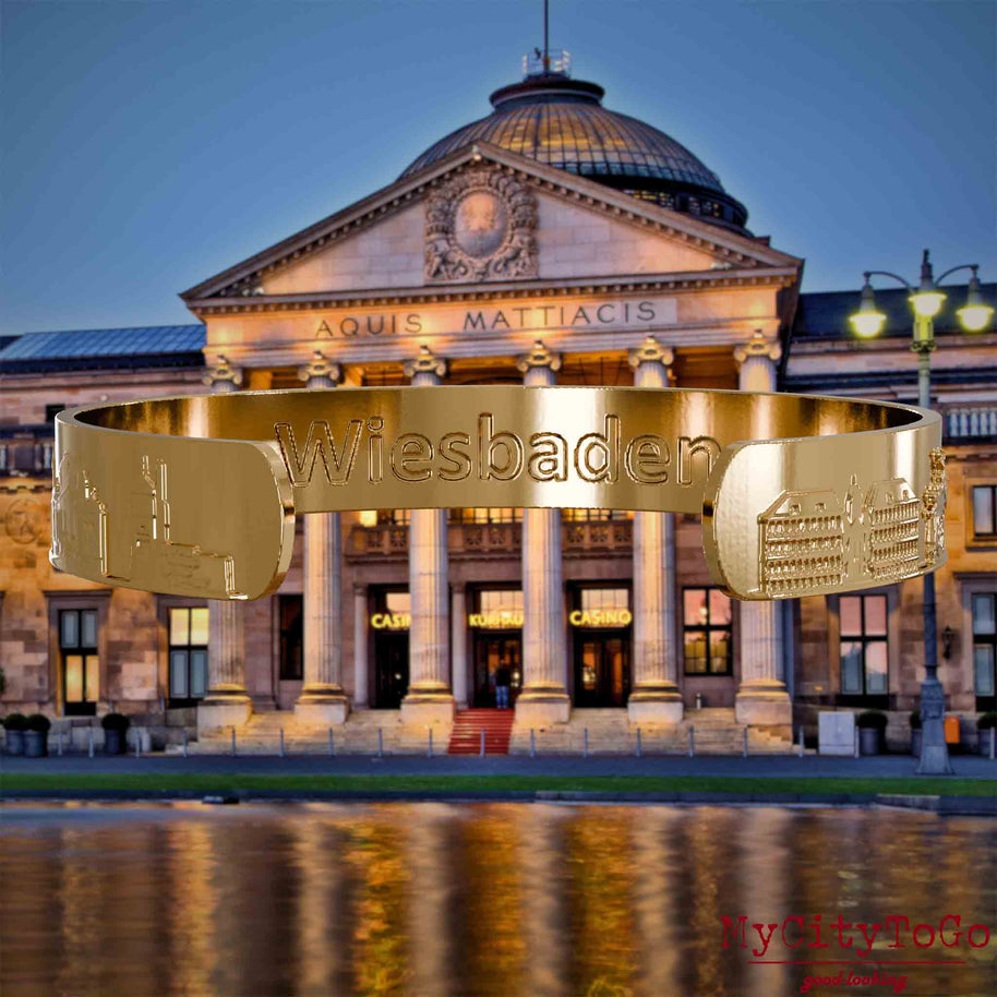 Innenseite eines vergoldeten Armreifs mit Reliefs bekannter Sehenswürdigkeiten der Stadt Wiesbaden und einer Gravur des Stadtnamens. Den Hintergrund bildet eine Ansicht der Stadt Wiesbaden.