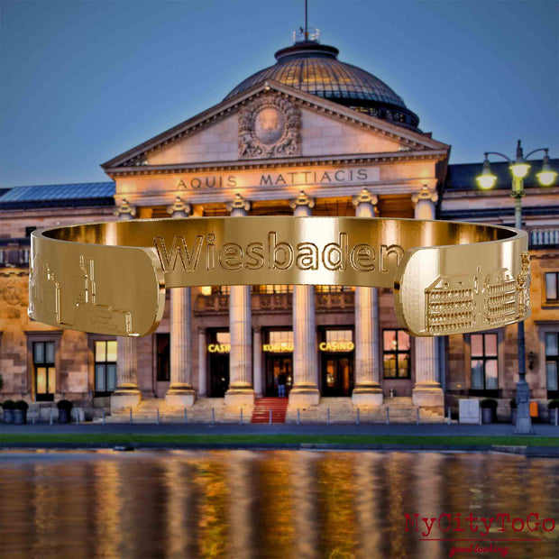 Innenseite eines vergoldeten Armreifs mit Reliefs bekannter Sehenswürdigkeiten der Stadt Wiesbaden und einer Gravur des Stadtnamens. Den Hintergrund bildet eine Ansicht der Stadt Wiesbaden.