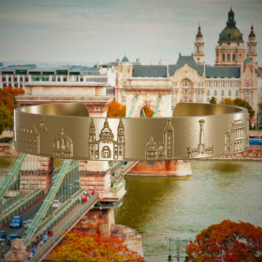 Armreif aus vergoldetem Edelstahl mit Reliefs bekannter Sehenswürdigkeiten der Stadt Budapest. Den Hintergrund bildet eine Ansicht der Stadt Budapest.