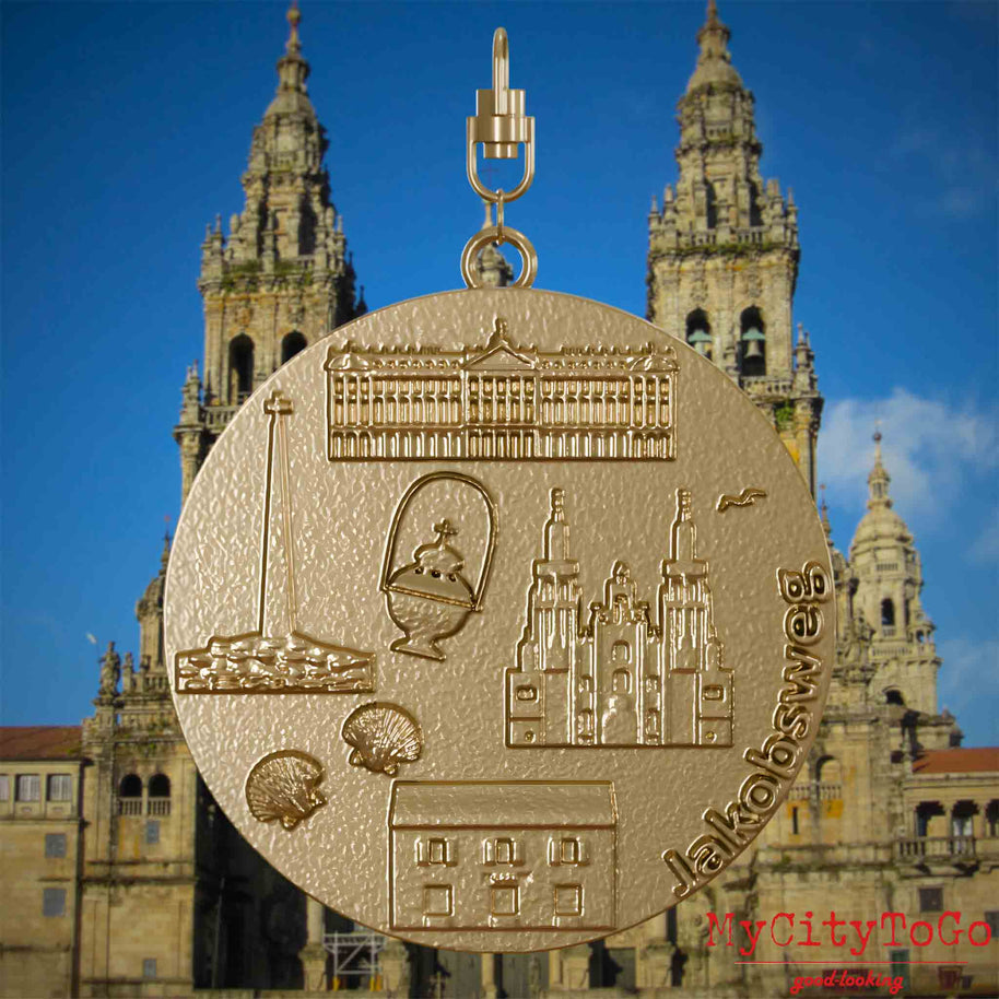 Halskette mit Drehwirbel und Rückseite eines Schmuckanhängers aus vergoldetem Edelstahl, auf dem Reliefs bekannter Sehenswürdigkeiten des Jakobswegs abgebildet sind. Den Hintergrund bildet eine Ansicht der Stadt Santiago de Compostela.
