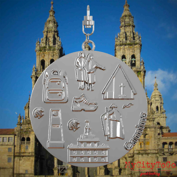 Halskette mit Drehwirbel und Vorderseite eines silbernen Schmuckanhängers, auf dem Reliefs bekannter Sehenswürdigkeiten des Jakobswegs abgebildet sind. Den Hintergrund bildet eine Ansicht der Stadt Santiago de Compostela.
