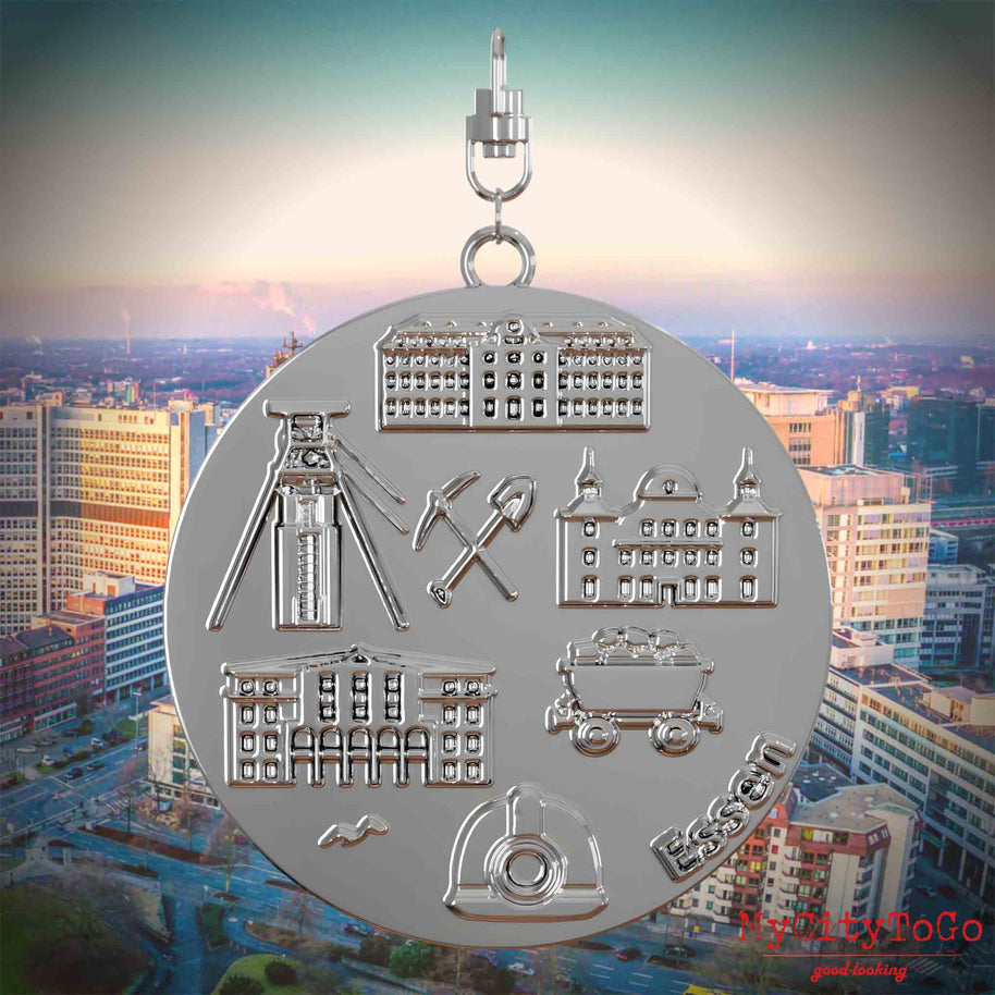 Halskette mit Drehwirbel und Rückseite eines silbernen Schmuckanhängers, auf dem Reliefs bekannter Sehenswürdigkeiten der Stadt Essen abgebildet sind. Den Hintergrund bildet eine Ansicht der Stadt Essen.