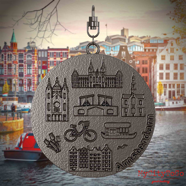 Halskette mit Drehwirbel und Vorderseite eines Schmuckanhängers aus Titan, auf dem Reliefs bekannter Sehenswürdigkeiten der Stadt Amsterdam abgebildet sind. Den Hintergrund bildet eine Ansicht der Stadt Amsterdam.
