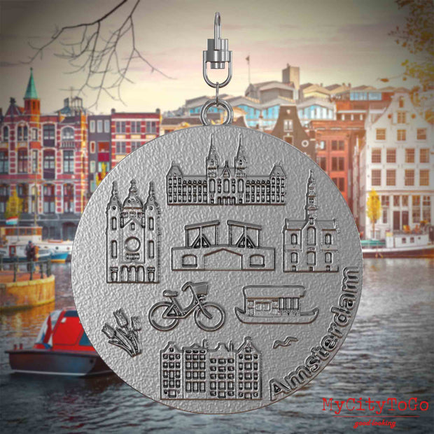 Halskette mit Drehwirbel und Vorderseite eines Schmuckanhängers aus Edelstahl, auf dem Reliefs bekannter Sehenswürdigkeiten der Stadt Amsterdam abgebildet sind. Den Hintergrund bildet eine Ansicht der Stadt Amsterdam.
