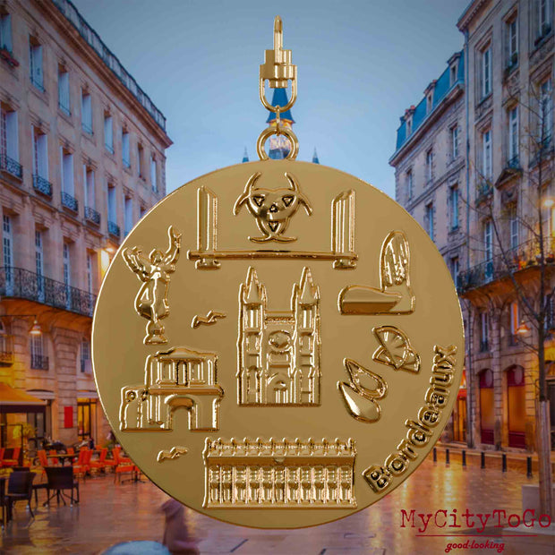 Halskette mit Drehwirbel und Vorderseite eines vergoldeten Schmuckanhängers, auf dem Reliefs bekannter Sehenswürdigkeiten der Stadt Bordeaux abgebildet sind. Den Hintergrund bildet eine Ansicht der Stadt Bordeaux.

