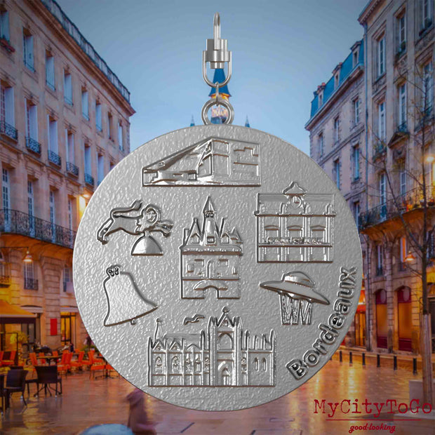 Halskette mit Drehwirbel und Rückseite eines Schmuckanhängers aus Edelstahl, auf dem Reliefs bekannter Sehenswürdigkeiten der Stadt Bordeaux abgebildet sind. Den Hintergrund bildet eine Ansicht der Stadt Bordeaux.

