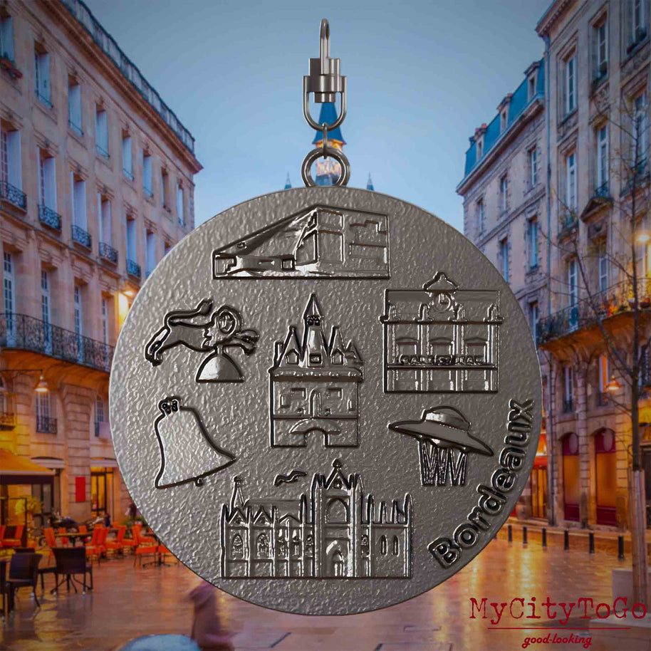 Halskette mit Drehwirbel und Rückseite eines Schmuckanhängers aus Titan, auf dem Reliefs bekannter Sehenswürdigkeiten der Stadt Bordeaux abgebildet sind. Den Hintergrund bildet eine Ansicht der Stadt Bordeaux.
