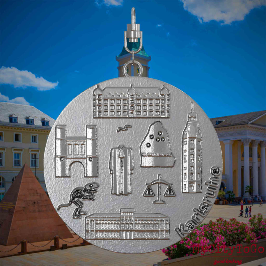 Halskette mit Drehwirbel und Rückseite eines Schmuckanhängers aus Edelstahl, auf dem Reliefs bekannter Sehenswürdigkeiten der Stadt Karlsruhe abgebildet sind. Den Hintergrund bildet eine Ansicht der Stadt Karlsruhe .
