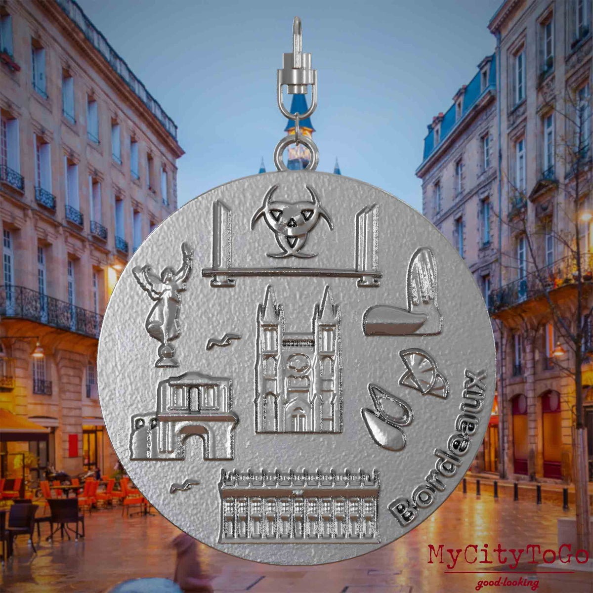 Halskette mit Drehwirbel und Vorderseite eines Schmuckanhängers aus Edelstahl, auf dem Reliefs bekannter Sehenswürdigkeiten der Stadt Bordeaux abgebildet sind. Den Hintergrund bildet eine Ansicht der Stadt Bordeaux.
