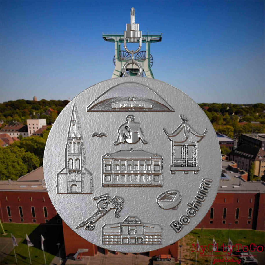Halskette mit Drehwirbel und Vorderseite eines Schmuckanhängers aus Edelstahl, auf dem Reliefs bekannter Sehenswürdigkeiten der Stadt Bochum abgebildet sind. Den Hintergrund bildet eine Ansicht der Stadt Bochum .

