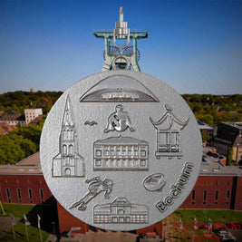 Halskette mit Drehwirbel und Vorderseite eines Schmuckanhängers aus Edelstahl, auf dem Reliefs bekannter Sehenswürdigkeiten der Stadt Bochum abgebildet sind. Den Hintergrund bildet eine Ansicht der Stadt Bochum .
