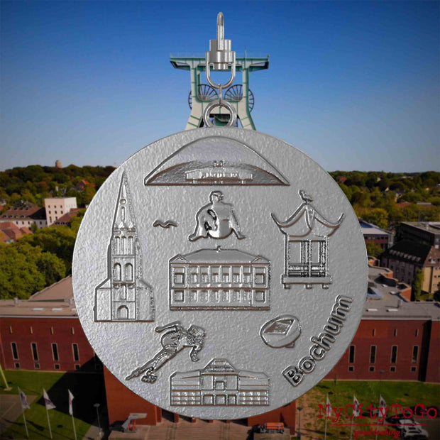 Halskette mit Drehwirbel und Vorderseite eines Schmuckanhängers aus Edelstahl, auf dem Reliefs bekannter Sehenswürdigkeiten der Stadt Bochum abgebildet sind. Den Hintergrund bildet eine Ansicht der Stadt Bochum .
