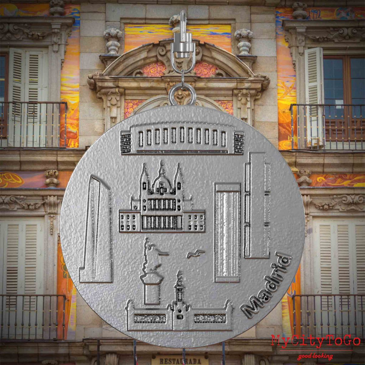 Halskette mit Drehwirbel und Rückseite eines Schmuckanhängers aus Edelstahl, auf dem Reliefs bekannter Sehenswürdigkeiten der Stadt Madrid abgebildet sind. Den Hintergrund bildet eine Ansicht der Stadt Madrid.
