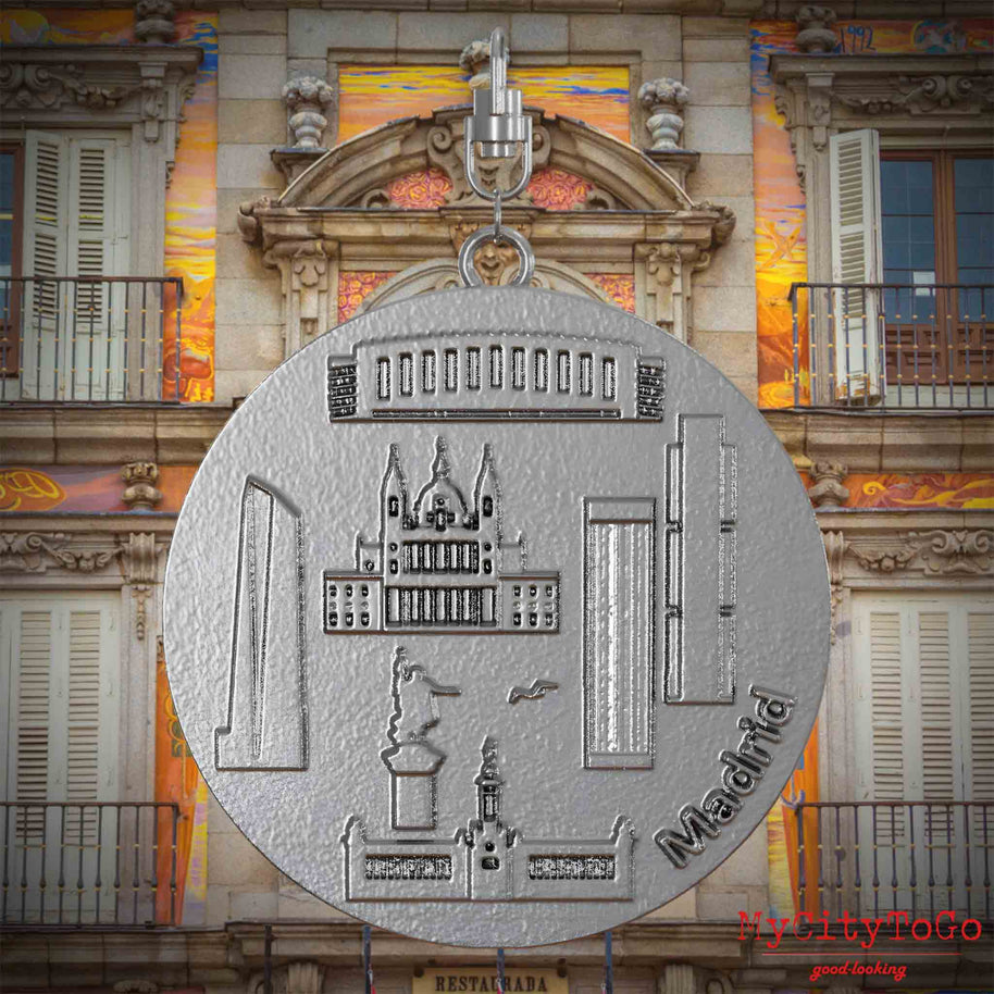 Halskette mit Drehwirbel und Rückseite eines Schmuckanhängers aus Edelstahl, auf dem Reliefs bekannter Sehenswürdigkeiten der Stadt Madrid abgebildet sind. Den Hintergrund bildet eine Ansicht der Stadt Madrid.
