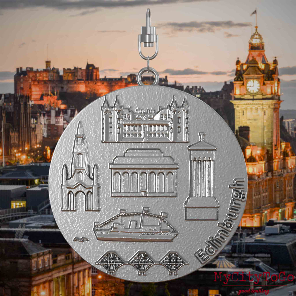 Halskette mit Drehwirbel und Rückseite eines Schmuckanhängers aus Edelstahl, auf dem Reliefs bekannter Sehenswürdigkeiten der Stadt Edinburgh abgebildet sind. Den Hintergrund bildet eine Ansicht der Stadt Edinburgh.

