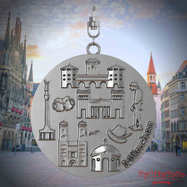 Halskette mit Drehwirbel und Vorderseite eines silbernen Schmuckanhängers, auf dem Reliefs bekannter Sehenswürdigkeiten der Stadt Muenchen abgebildet sind. Den Hintergrund bildet eine Ansicht der Stadt Muenchen.