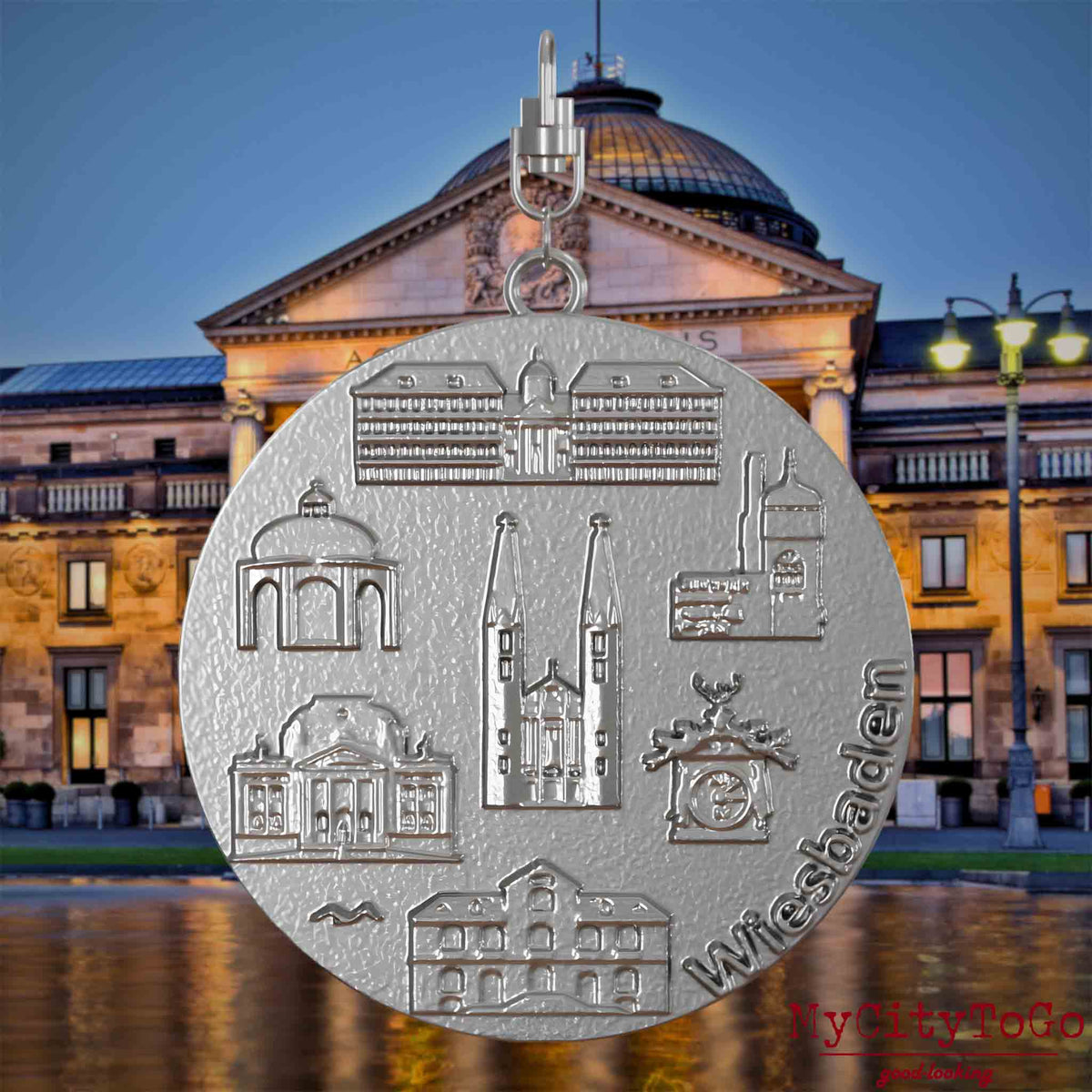 Halskette mit Drehwirbel und Rückseite eines Schmuckanhängers aus Edelstahl, auf dem Reliefs bekannter Sehenswürdigkeiten der Stadt Wiesbaden abgebildet sind. Den Hintergrund bildet eine Ansicht der Stadt Wiesbaden.

