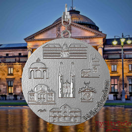 Halskette mit Drehwirbel und Rückseite eines Schmuckanhängers aus Edelstahl, auf dem Reliefs bekannter Sehenswürdigkeiten der Stadt Wiesbaden abgebildet sind. Den Hintergrund bildet eine Ansicht der Stadt Wiesbaden.
