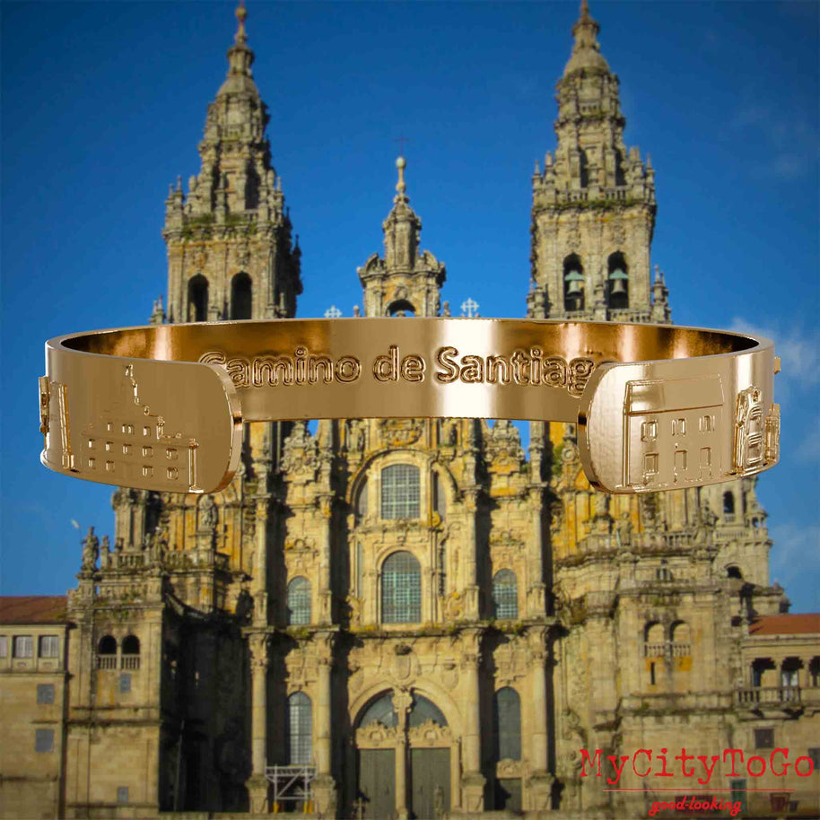 Innenseite eines vergoldeten Armreifs mit Reliefs bekannter Sehenswürdigkeiten und Elemente des Jakobsweges und der Gravur Camino de Santiago. Den Hintergrund bildet die Kathedrale von Santiago de Compostela.