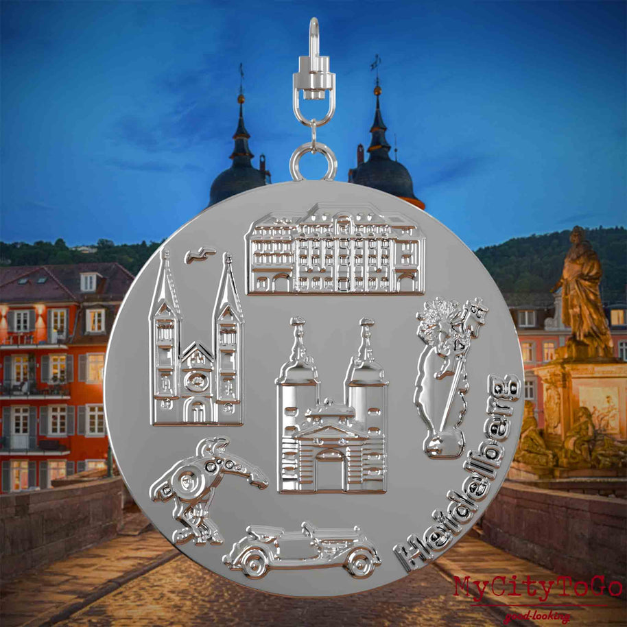 Halskette mit Drehwirbel und Rückseite eines silbernen Schmuckanhängers, auf dem Reliefs bekannter Sehenswürdigkeiten der Stadt Heidelberg abgebildet sind. Den Hintergrund bildet eine Ansicht der Stadt Heidelberg .
