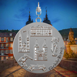 Halskette mit Drehwirbel und Rückseite eines silbernen Schmuckanhängers, auf dem Reliefs bekannter Sehenswürdigkeiten der Stadt Heidelberg abgebildet sind. Den Hintergrund bildet eine Ansicht der Stadt Heidelberg .

