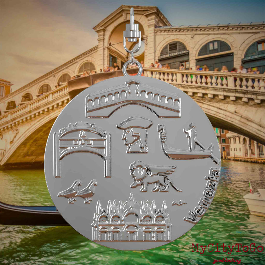 Halskette mit Drehwirbel und Vorderseite eines silbernen Schmuckanhängers, auf dem Reliefs bekannter Sehenswürdigkeiten der Stadt Venedig abgebildet sind. Den Hintergrund bildet eine Ansicht der Stadt Venedig.
