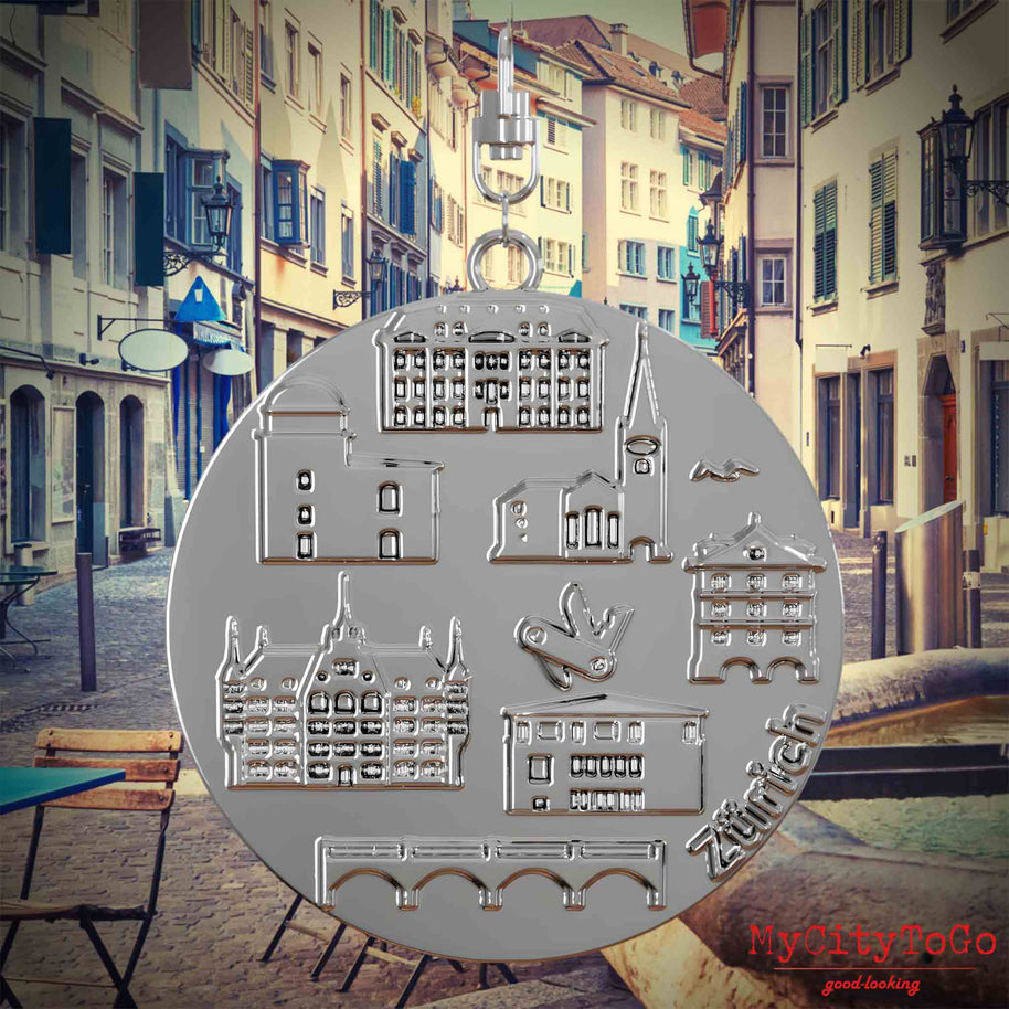 Halskette mit Drehwirbel und Rückseite eines silbernen Schmuckanhängers, auf dem Reliefs bekannter Sehenswürdigkeiten der Stadt Zuerich abgebildet sind. Den Hintergrund bildet eine Ansicht der Stadt Zuerich.
