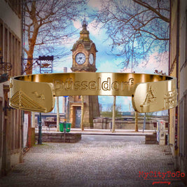 Innenseite eines vergoldeten Armreifs mit Reliefs bekannter Sehenswürdigkeiten der Stadt Duesseldorf und einer Gravur des Stadtnamens. Den Hintergrund bildet eine Ansicht der Stadt Duesseldorf.