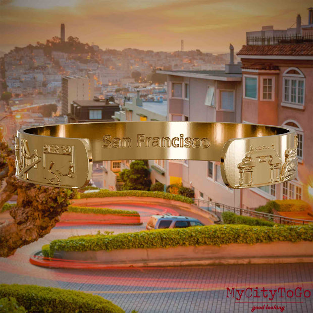 Innenseite eines vergoldeten Armreifs mit Reliefs bekannter Sehenswürdigkeiten der Stadt San Francisco und einer Gravur des Stadtnamens. Den Hintergrund bildet eine Ansicht der Stadt San Francisco.