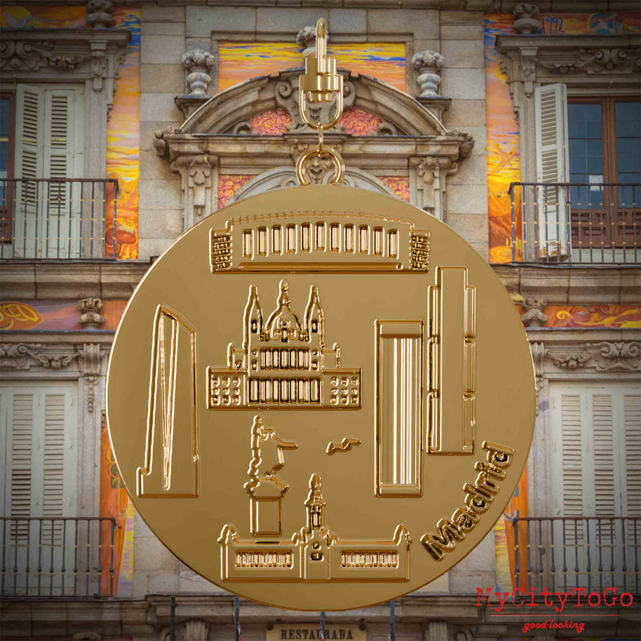 Halskette mit Drehwirbel und Rückseite eines vergoldeten Schmuckanhängers, auf dem Reliefs bekannter Sehenswürdigkeiten der Stadt Madrid abgebildet sind. Den Hintergrund bildet eine Ansicht der Stadt Madrid.
