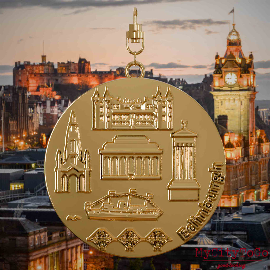 Halskette mit Drehwirbel und Rückseite eines vergoldeten Schmuckanhängers, auf dem Reliefs bekannter Sehenswürdigkeiten der Stadt Edinburgh abgebildet sind. Den Hintergrund bildet eine Ansicht der Stadt Edinburgh.
