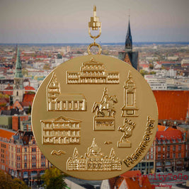 Halskette mit Drehwirbel und Rückseite eines vergoldeten Schmuckanhängers, auf dem Reliefs bekannter Sehenswürdigkeiten der Stadt Hannover abgebildet sind. Den Hintergrund bildet eine Ansicht der Stadt Hannover .
