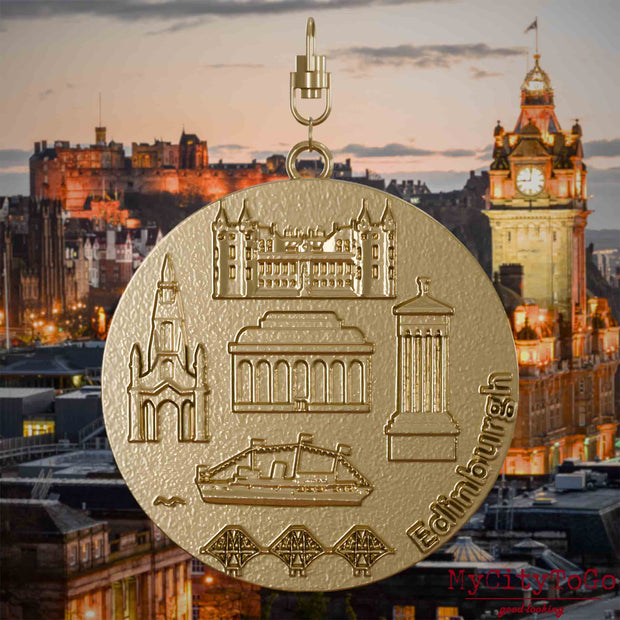 Halskette mit Drehwirbel und Rückseite eines Schmuckanhängers aus vergoldetem Edelstahl, auf dem Reliefs bekannter Sehenswürdigkeiten der Stadt Edinburgh abgebildet sind. Den Hintergrund bildet eine Ansicht der Stadt Edinburgh.
