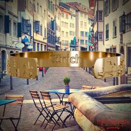 Innenseite eines vergoldeten Armreifs mit Reliefs bekannter Sehenswürdigkeiten der Stadt Zuerich und einer Gravur des Stadtnamens . Den Hintergrund bildet eine Ansicht der Stadt Zuerich.