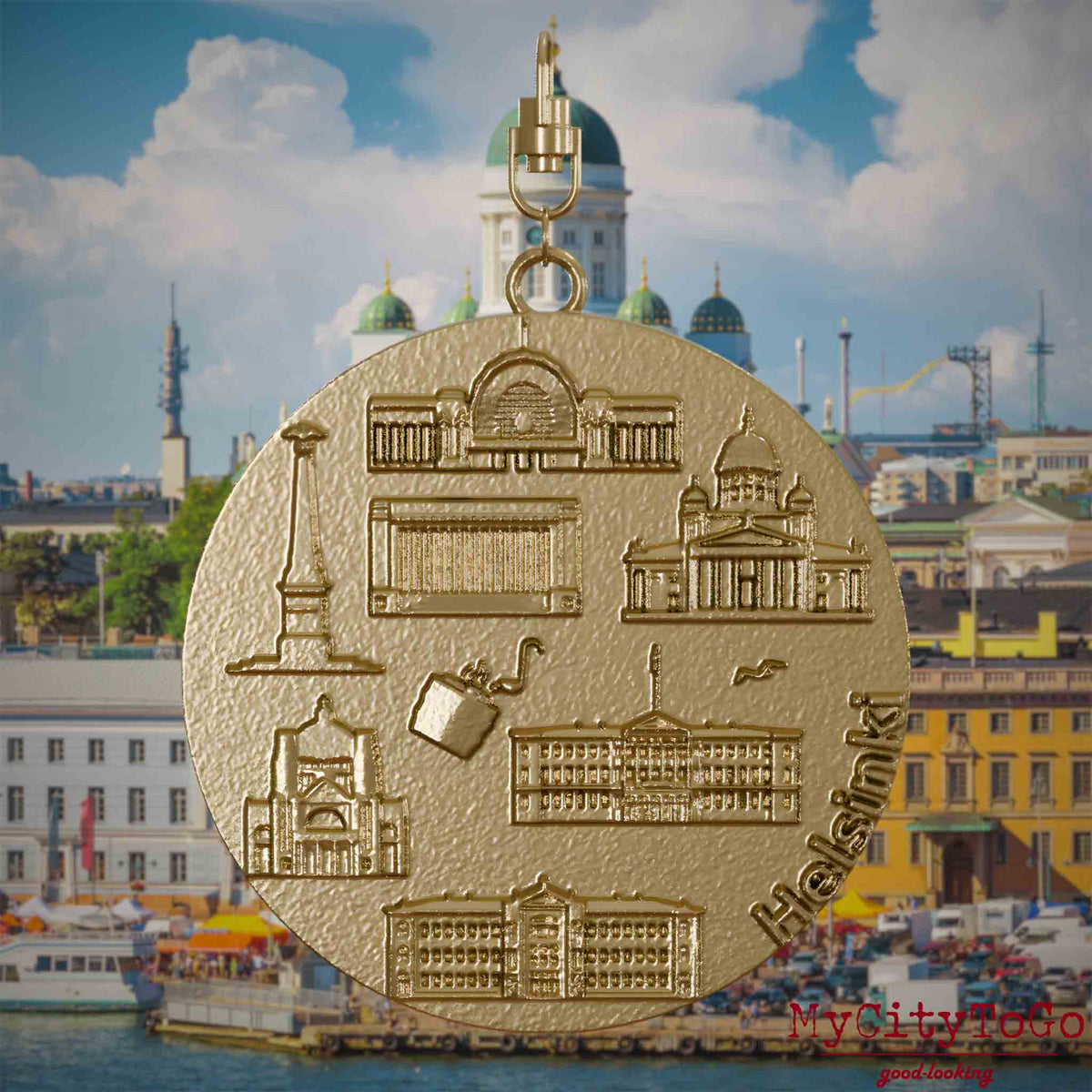 Halskette mit Drehwirbel und Vorderseite eines Schmuckanhängers aus vergoldetem Edelstahl, auf dem Reliefs bekannter Sehenswürdigkeiten der Stadt Helsinki abgebildet sind. Den Hintergrund bildet eine Ansicht der Stadt Helsinki.
