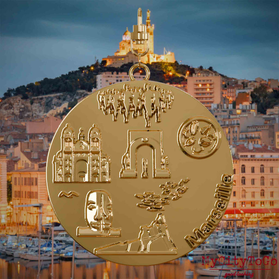 Halskette mit Drehwirbel und Rückseite eines vergoldeten Schmuckanhängers, auf dem Reliefs bekannter Sehenswürdigkeiten der Stadt Marseille abgebildet sind. Den Hintergrund bildet eine Ansicht der Stadt Marseille.
