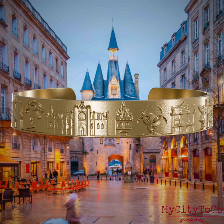 Armreif aus vergoldetem Edelstahl mit Reliefs bekannter Sehenswürdigkeiten der Stadt Bordeaux. Den Hintergrund bildet eine Ansicht der Stadt Bordeaux.
