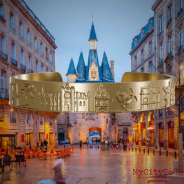 Armreif aus vergoldetem Edelstahl mit Reliefs bekannter Sehenswürdigkeiten der Stadt Bordeaux. Den Hintergrund bildet eine Ansicht der Stadt Bordeaux.