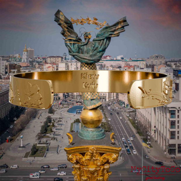 Innenseite eines vergoldeten Armreifs mit Reliefs bekannter Sehenswürdigkeiten der Stadt Kiew und einer Gravur des Stadtnamens. Den Hintergrund bildet eine Ansicht der Stadt Kiew.