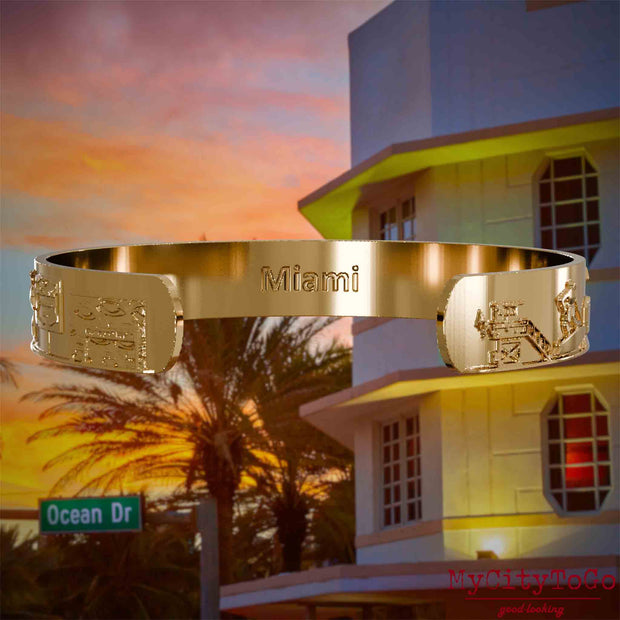 Innenseite eines vergoldeten Armreifs mit Reliefs bekannter Sehenswürdigkeiten der Stadt Miami und einer Gravur des Stadtnamens. Den Hintergrund bildet eine Ansicht der Stadt Miami.