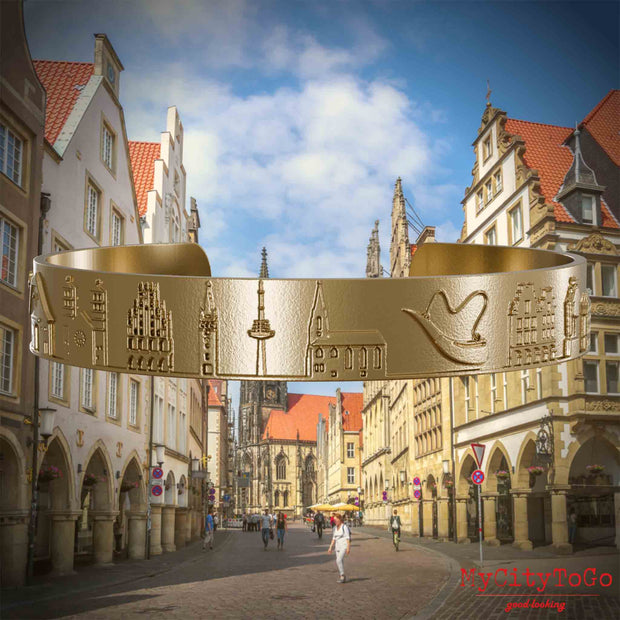 Armreif aus vergoldetem Edelstahl mit Reliefs bekannter Sehenswürdigkeiten der Stadt Muenster. Den Hintergrund bildet eine Ansicht der Stadt Muenster.