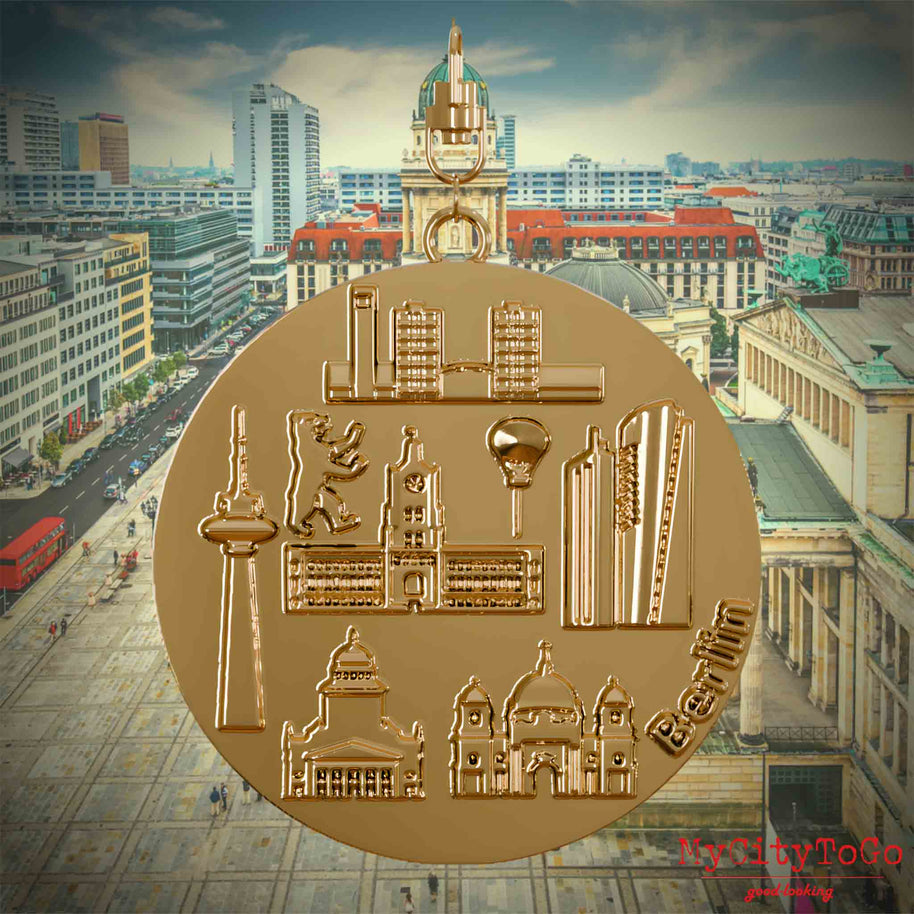 Halskette mit Drehwirbel und Vorderseite eines vergoldeten Schmuckanhängers, auf dem Reliefs bekannter Sehenswürdigkeiten der Stadt Berlin abgebildet sind. Den Hintergrund bildet eine Ansicht der Stadt Berlin.