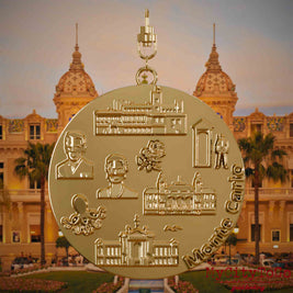 Halskette mit Drehwirbel und Rückseite eines vergoldeten Schmuckanhängers, auf dem Reliefs bekannter Sehenswürdigkeiten der Stadt Monte Carlo abgebildet sind. Den Hintergrund bildet eine Ansicht der Stadt Monte Carlo.
