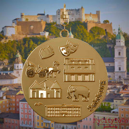 Halskette mit Drehwirbel und Rückseite eines vergoldeten Schmuckanhängers, auf dem Reliefs bekannter Sehenswürdigkeiten der Stadt Salzburg abgebildet sind. Den Hintergrund bildet eine Ansicht der Stadt Salzburg.
