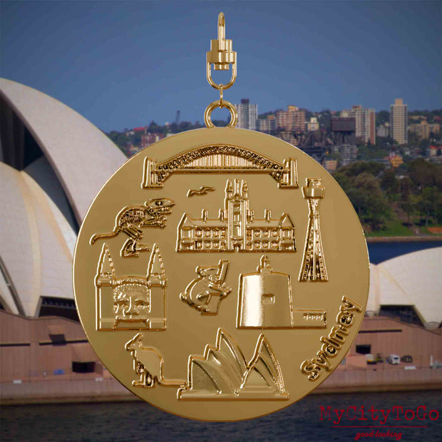 Halskette mit Drehwirbel und Vorderseite eines vergoldeten Schmuckanhängers, auf dem Reliefs bekannter Sehenswürdigkeiten der Stadt Sydney abgebildet sind. Den Hintergrund bildet eine Ansicht der Stadt Sydney.
