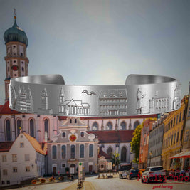 Armreif aus Edelstahl mit Reliefs bekannter Sehenswürdigkeiten der Stadt Augsburg. Den Hintergrund bildet eine Ansicht der Stadt Augsburg.