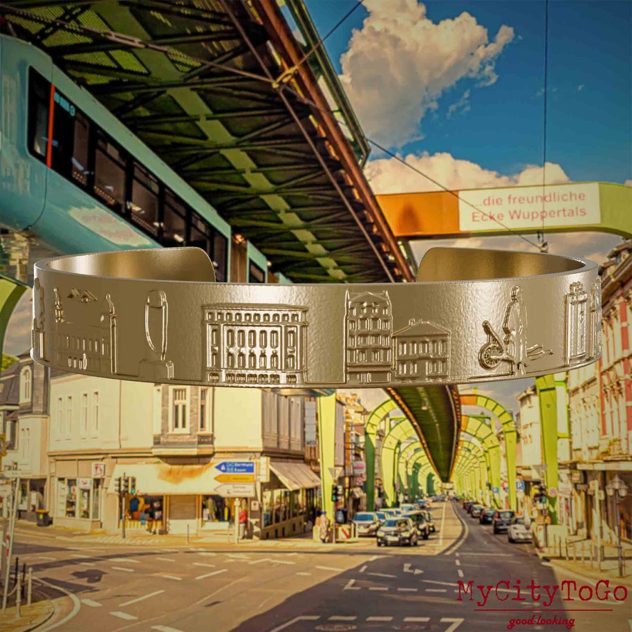 Armreif aus vergoldetem Edelstahl mit Reliefs bekannter Sehenswürdigkeiten der Stadt Wuppertal. Den Hintergrund bildet eine Ansicht  der Stadt Wuppertal.