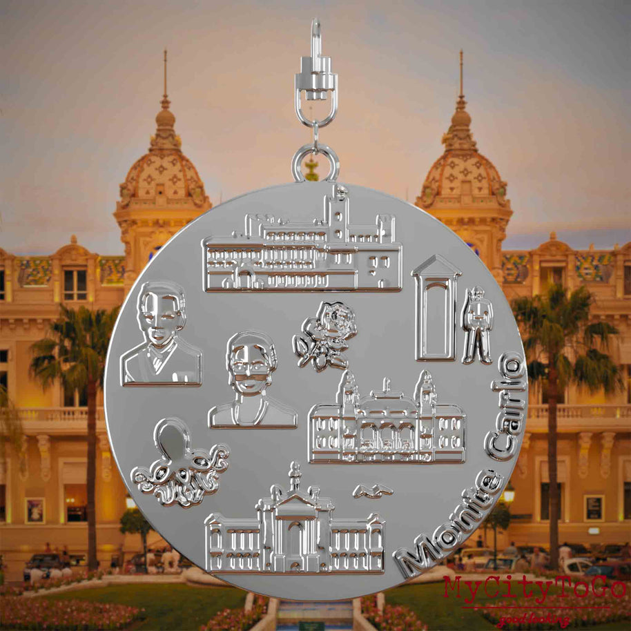 Halskette mit Drehwirbel und Rückseite eines silbernen Schmuckanhängers, auf dem Reliefs bekannter Sehenswürdigkeiten der Stadt Monte Carlo abgebildet sind. Den Hintergrund bildet eine Ansicht der Stadt Monte Carlo.
